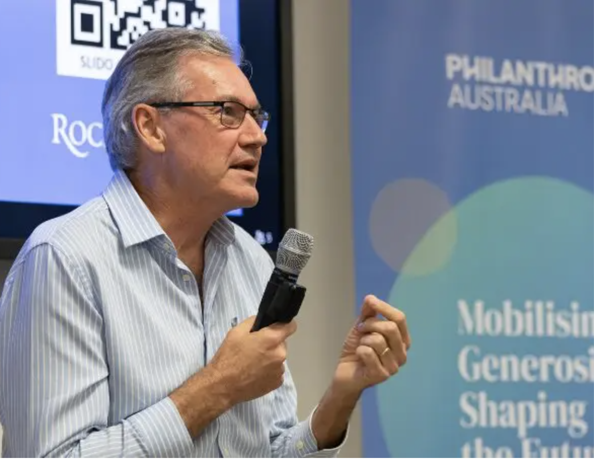 Darrell Wade PA Conference Darrell Wade speaking at Philanthropy Australia event holding microphone wearing blue button down shirt in front of blue signage