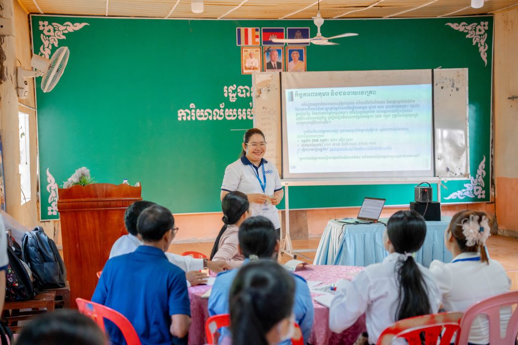 Teacher standing in front of classroom of children, smiling, in front of projector board