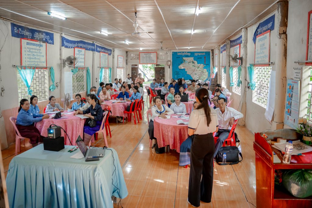 Teachers sitting at multiple round tables in a room with blue and pink table clothes, listening to a speaker standing with their back to the camera