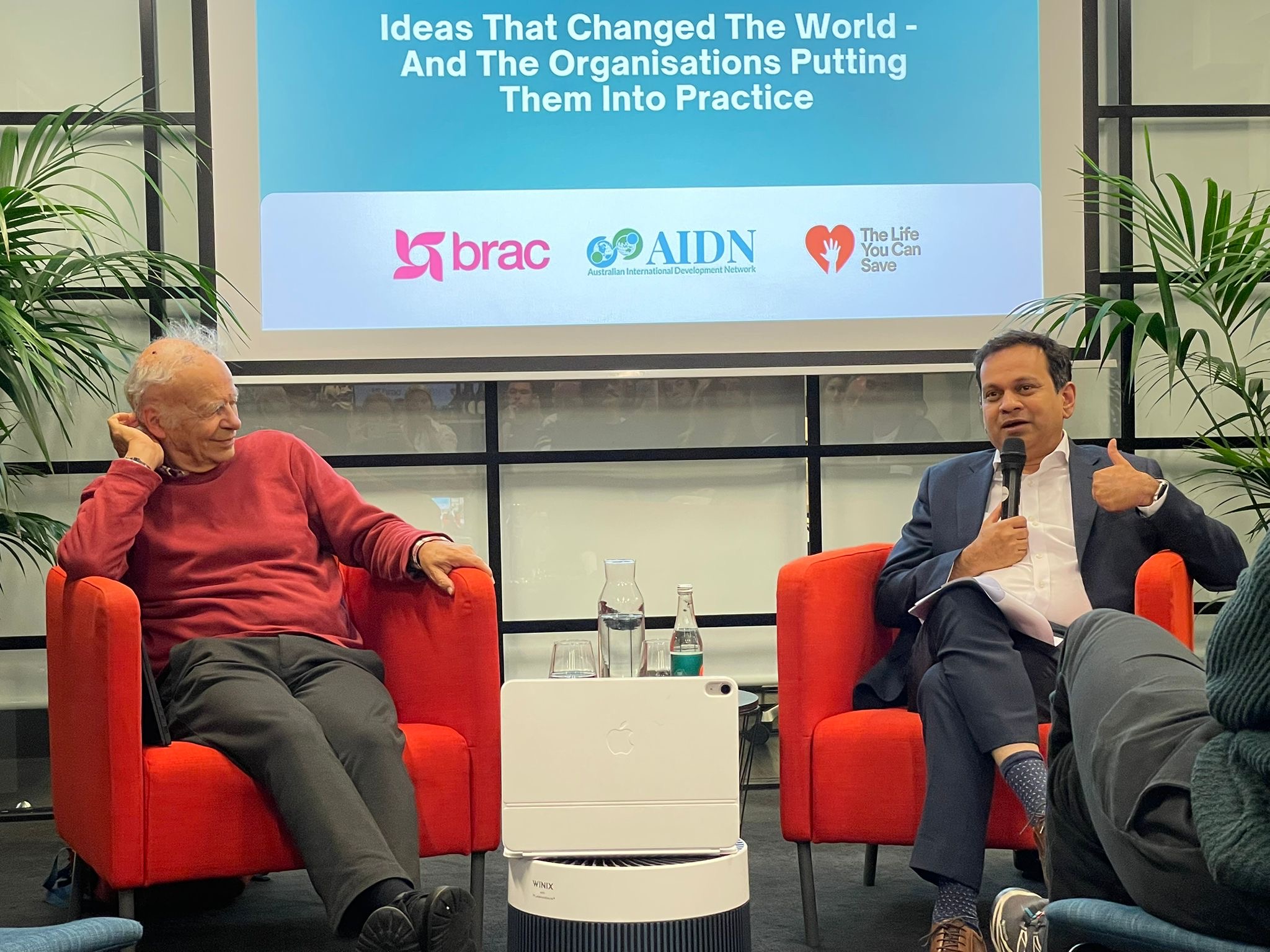 TLYCS/BRAC Ideas That Changed The World Event Two men sitting on stage in red chairs, one holding a microphone, in front of presentation screen