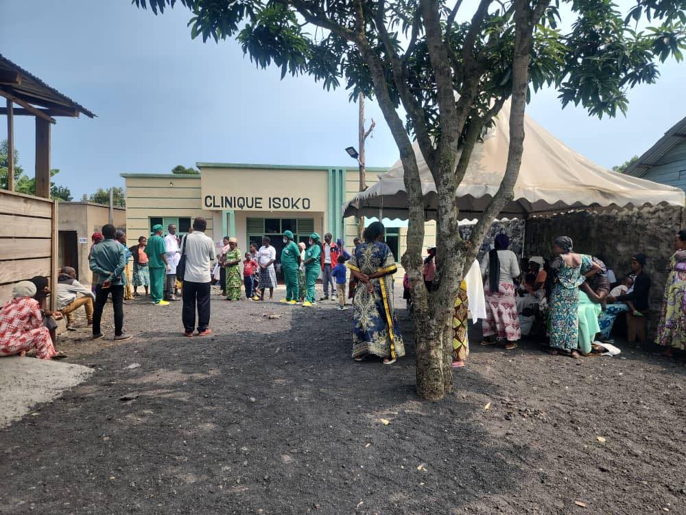 Crowd of healthcare workers and patients standing outside a clinic under a tree and tent, wearing PPE or colourful dresses