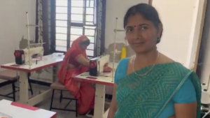 Woman wearing green traditional garment smiling at camera in foreground, with woman in red traditional garment sitting at sewing machine in background