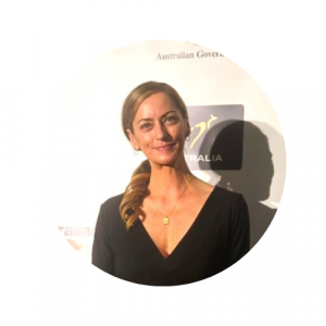 Sally Miles smiling at camera in front of Australian Aid back drop, with hair in low ponytail wearing black V-neck shirt