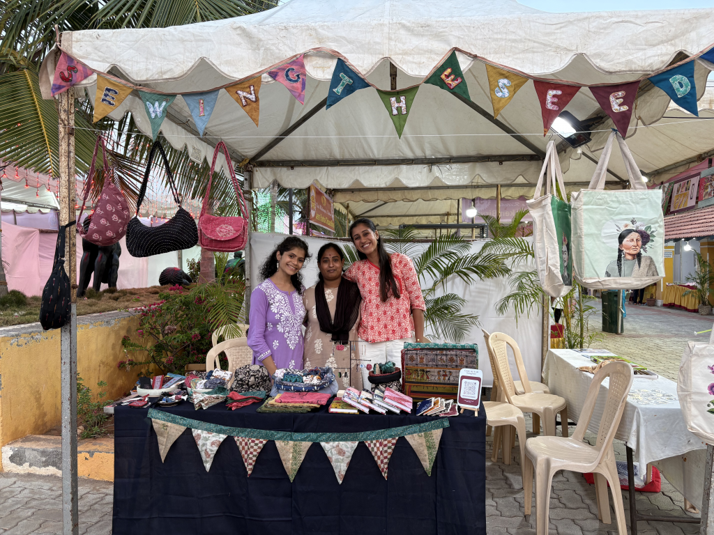 3 women standing under white tent decorated with hand-sewn bunting behind table selling sewn wares