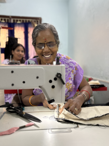 Woman wearing lilac clothes and glasses sitting behind a sewing machine working on a piece, smiling