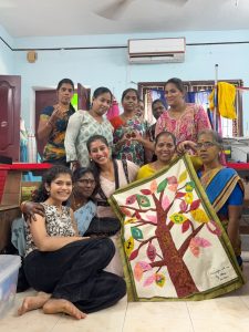 Women standing and sitting in a group, smiling at the camera, some holding a large quilt with a tree with colourful leaves on it